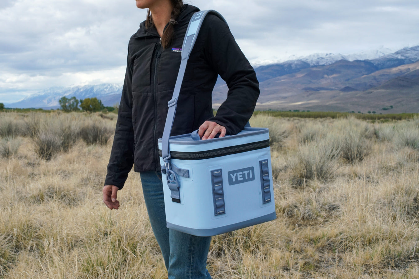 A person standing in an outdoor grassy field with mountains in the background carries a light blue YETI cooler bag with a gray strap.
